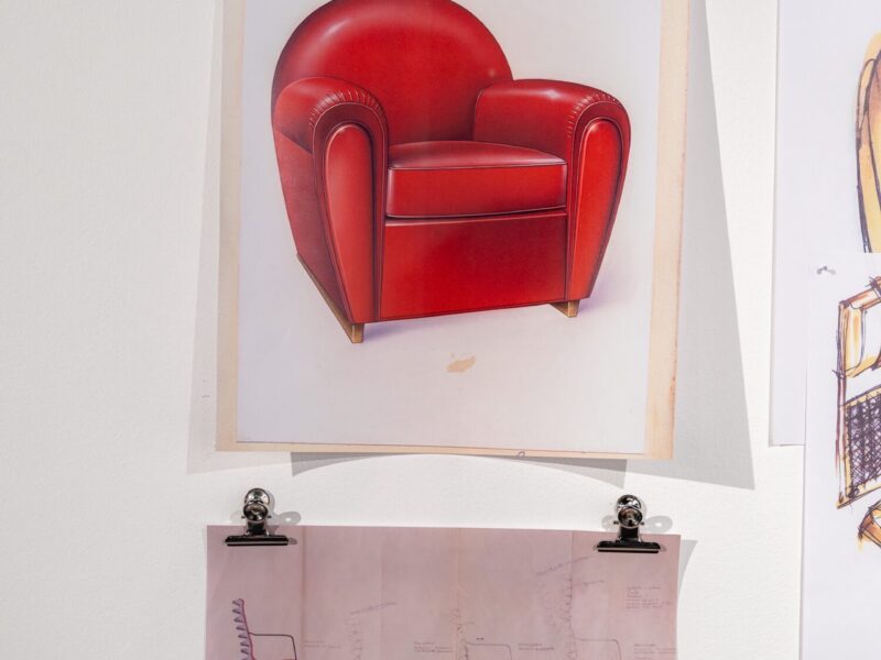 Tavola illustrata a colori di una poltrona in pelle rossa, esposta su un pannello con fermagli nella mostra Abitare l'automobile