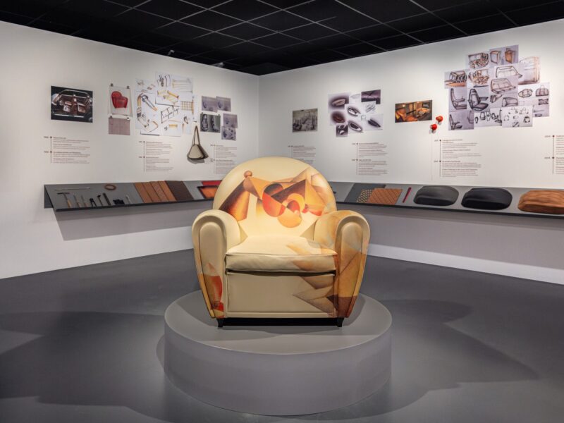 Poltrona con rivestimento a stampa astratta multicolore esposta su un piedistallo al centro della sala della mostra Abitare l'automobile, con bozzetti e pannelli informativi alle pareti