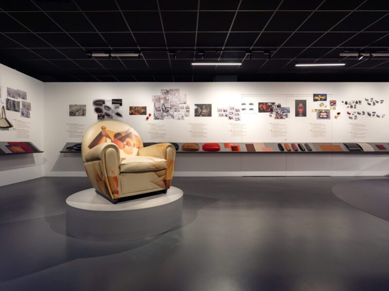 Vista d'insieme della sala della mostra Abitare l'automobile con la poltrona dalla fantasia astratta multicolore in primo piano e una lunga parete di bozzetti e pannelli espositivi sullo sfondo