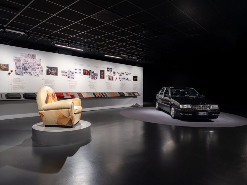 Sala della mostra Abitare l'automobile con la poltrona dalla fantasia astratta multicolore e un'auto d'epoca nera esposte su piedistalli, davanti a una parete di bozzetti di design
