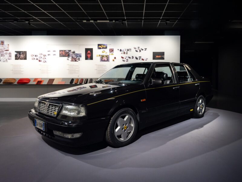 Auto d'epoca nera con filetto di rifinitura giallo esposta in sala nella mostra Abitare l'automobile, con una parete di bozzetti di design sullo sfondo