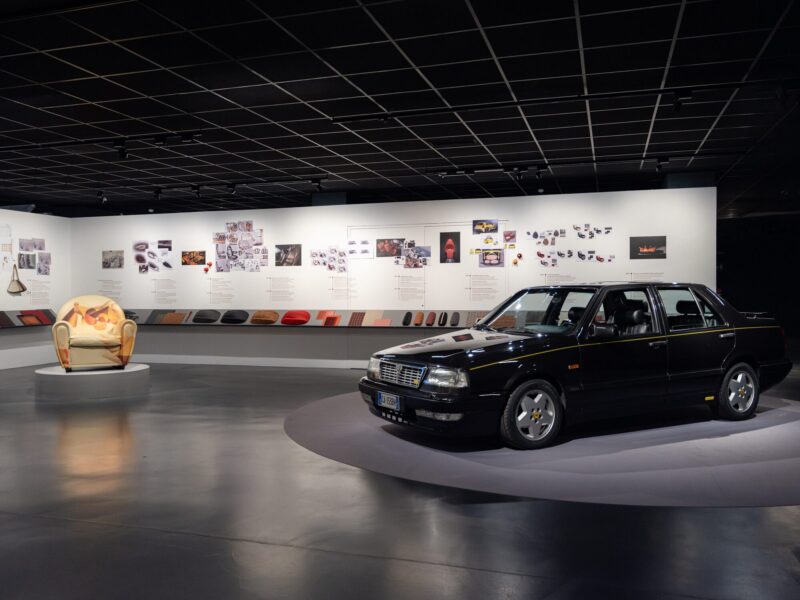 Vista panoramica della sala della mostra Abitare l'automobile con l'auto d'epoca nera in primo piano e la poltrona dalla fantasia astratta multicolore sullo sfondo, davanti a una parete di bozzetti