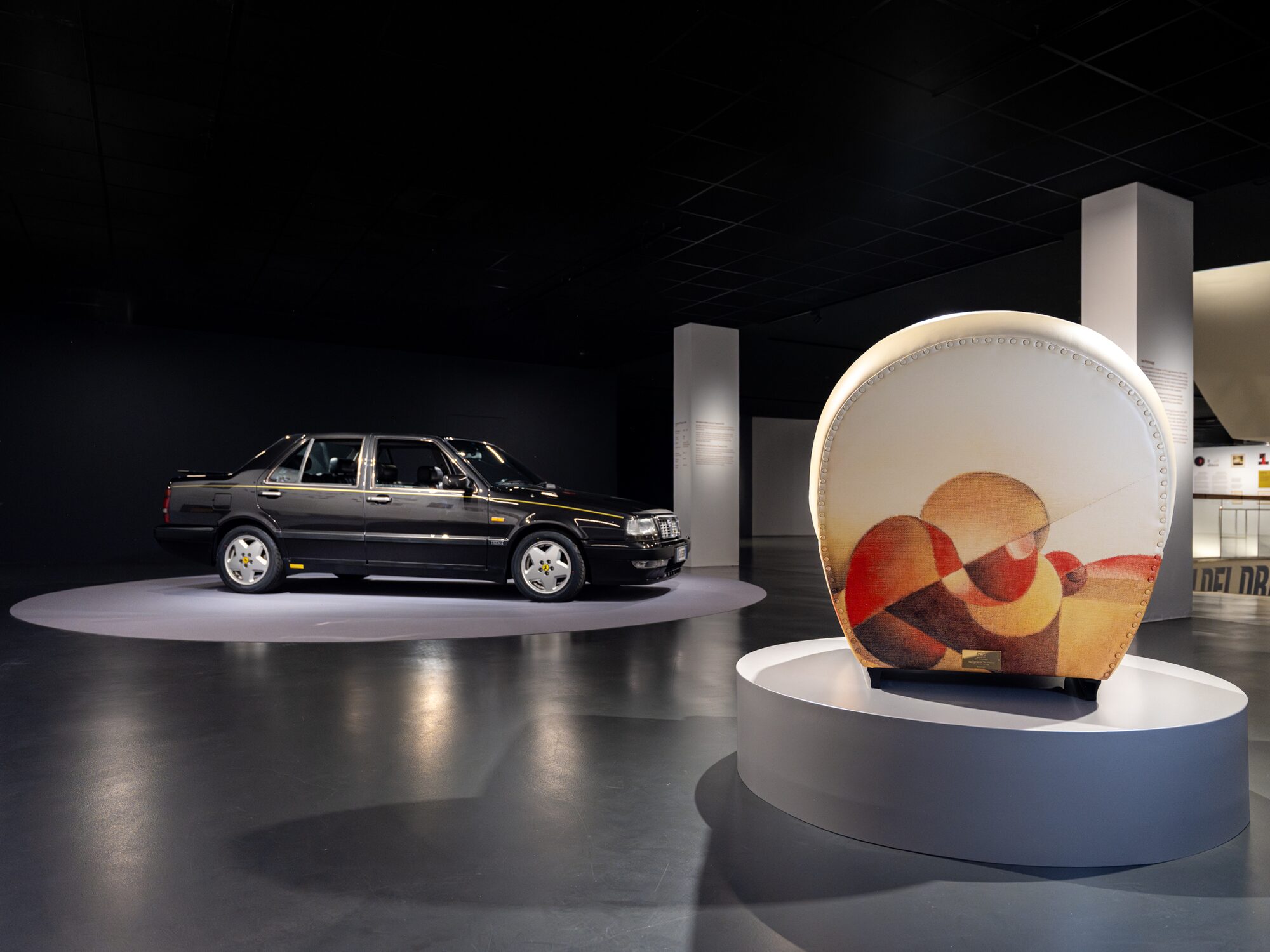 Schienale circolare di una poltrona dalla fantasia astratta multicolore in primo piano e auto d'epoca nera sullo sfondo, esposte nella mostra Abitare l'automobile