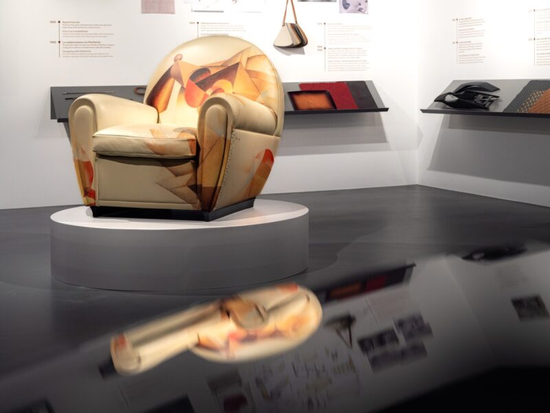 Poltrona vista di profilo con rivestimento a fantasia astratta multicolore esposta su un piedistallo, con campioni di materiali su una mensola sullo sfondo, mostra Abitare l'automobile