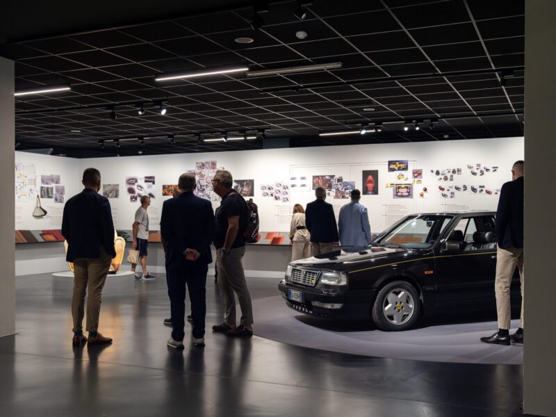 Visitatori osservano l'auto d'epoca nera e i pannelli espositivi durante la visita alla mostra Abitare l'automobile