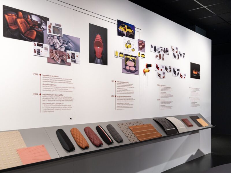 Parete cronologica della mostra Abitare l'automobile con immagini di collaborazioni automobilistiche e accessori, sopra una mensola con numerosi campioni di pelle e tessuto di diversi colori