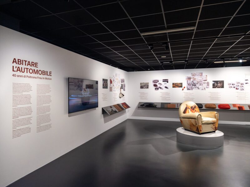 Ingresso della mostra Abitare l'automobile - 40 anni di Poltrona Frau In Motion, con testo introduttivo a parete, schermo video e la poltrona dalla fantasia astratta multicolore su piedistallo